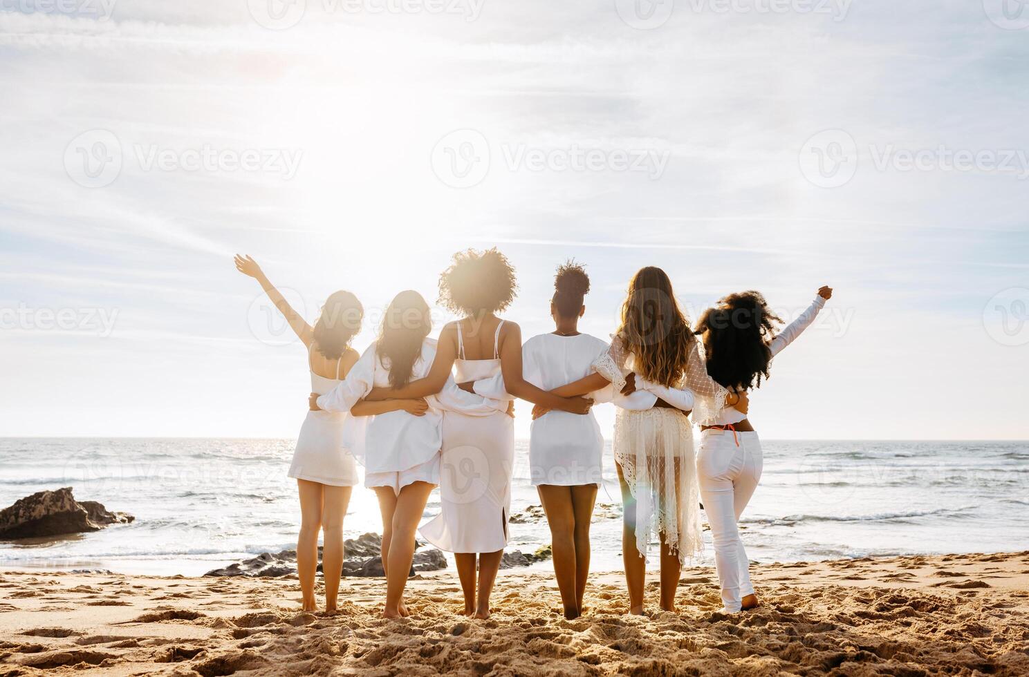 Bride's team. Back view of group of young diverse women posing and embracing by the seashore, enjoying sunset and beautiful ocean view. Hen party celebration photo