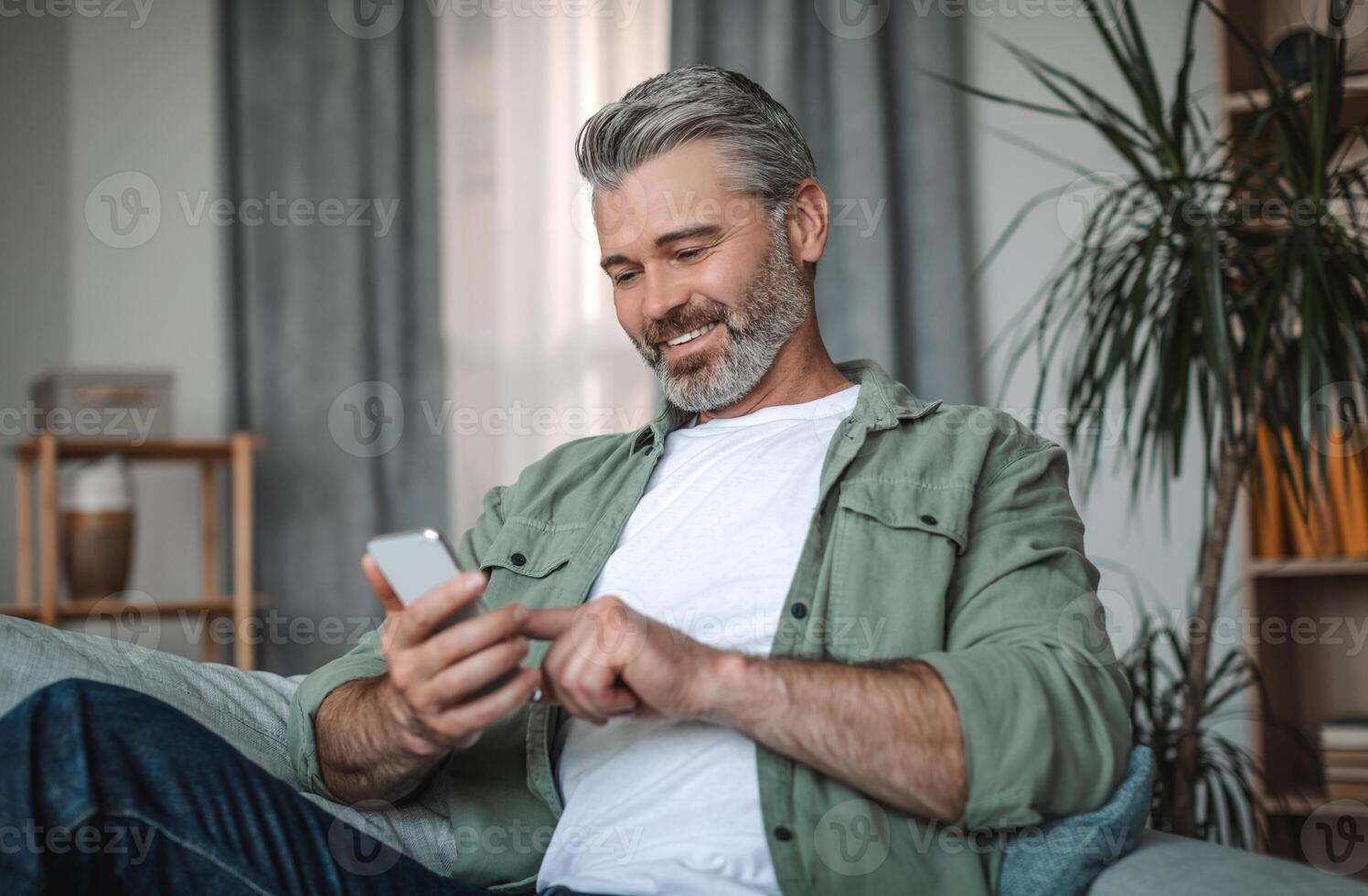 Cheerful elderly european man with beard typing on smartphone, enjoying social media chat in room interior. Social distance and new application, device for pensioner at home due covid-19 pandemic photo