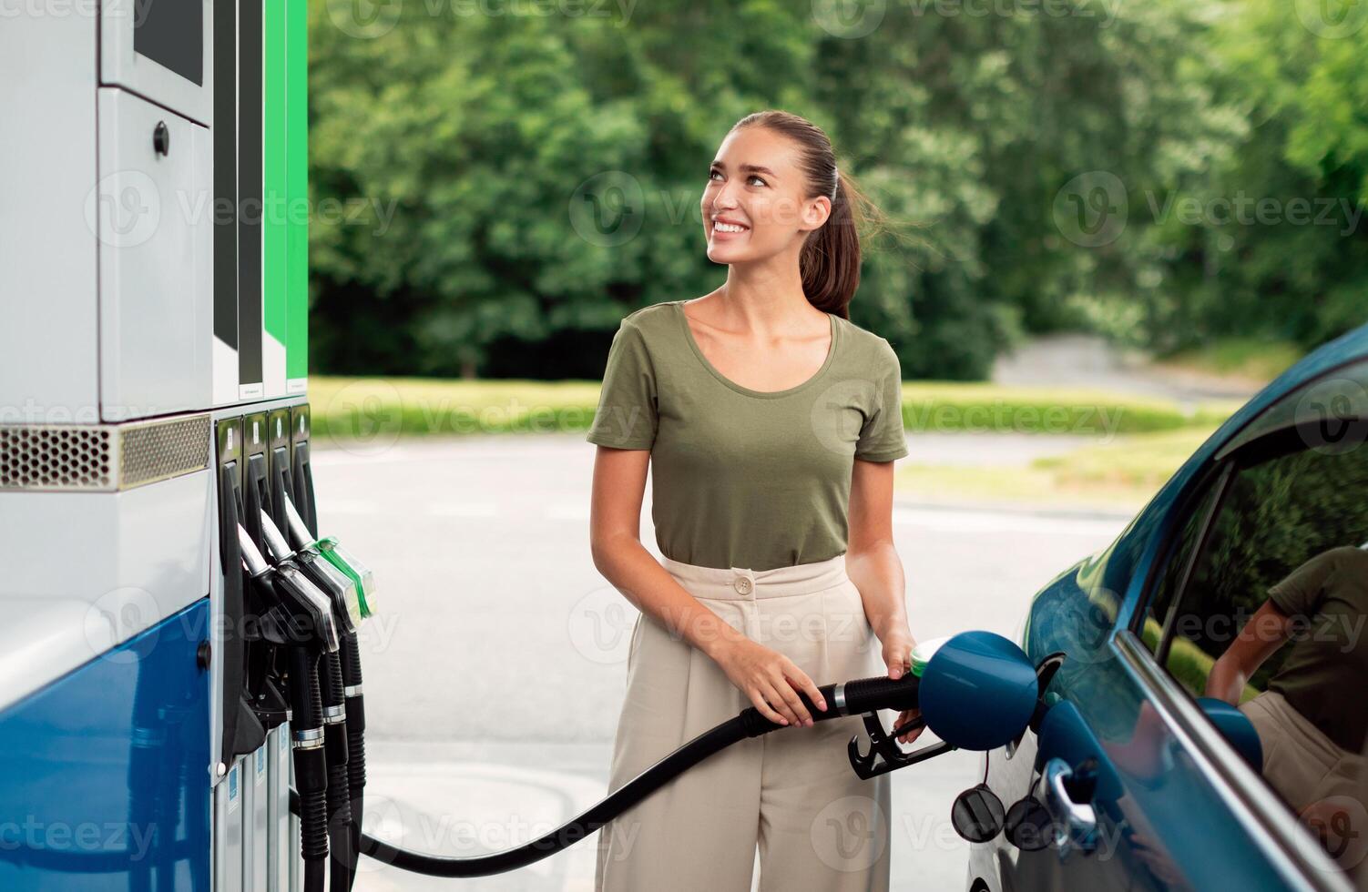 Happy Young Woman Refueling Her Car At Modern Gas Station Outdoor. Contented Driver Lady Standing Near Vehicle Holding Filling Gun Nozzle Filling Up Vehicle With Bio Fuel Near Petrol Dispenser photo