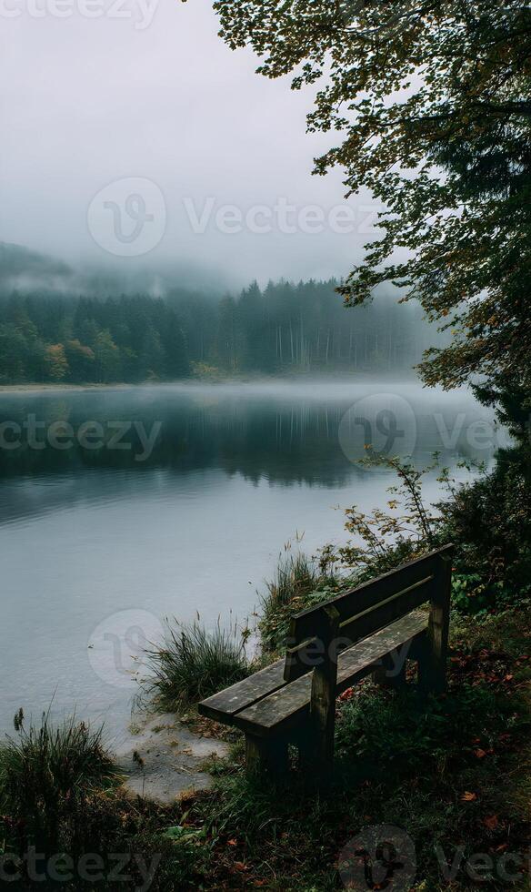 A tranquil scene unfolds with a wooden bench overlooking a misty lake and a serene forest photo