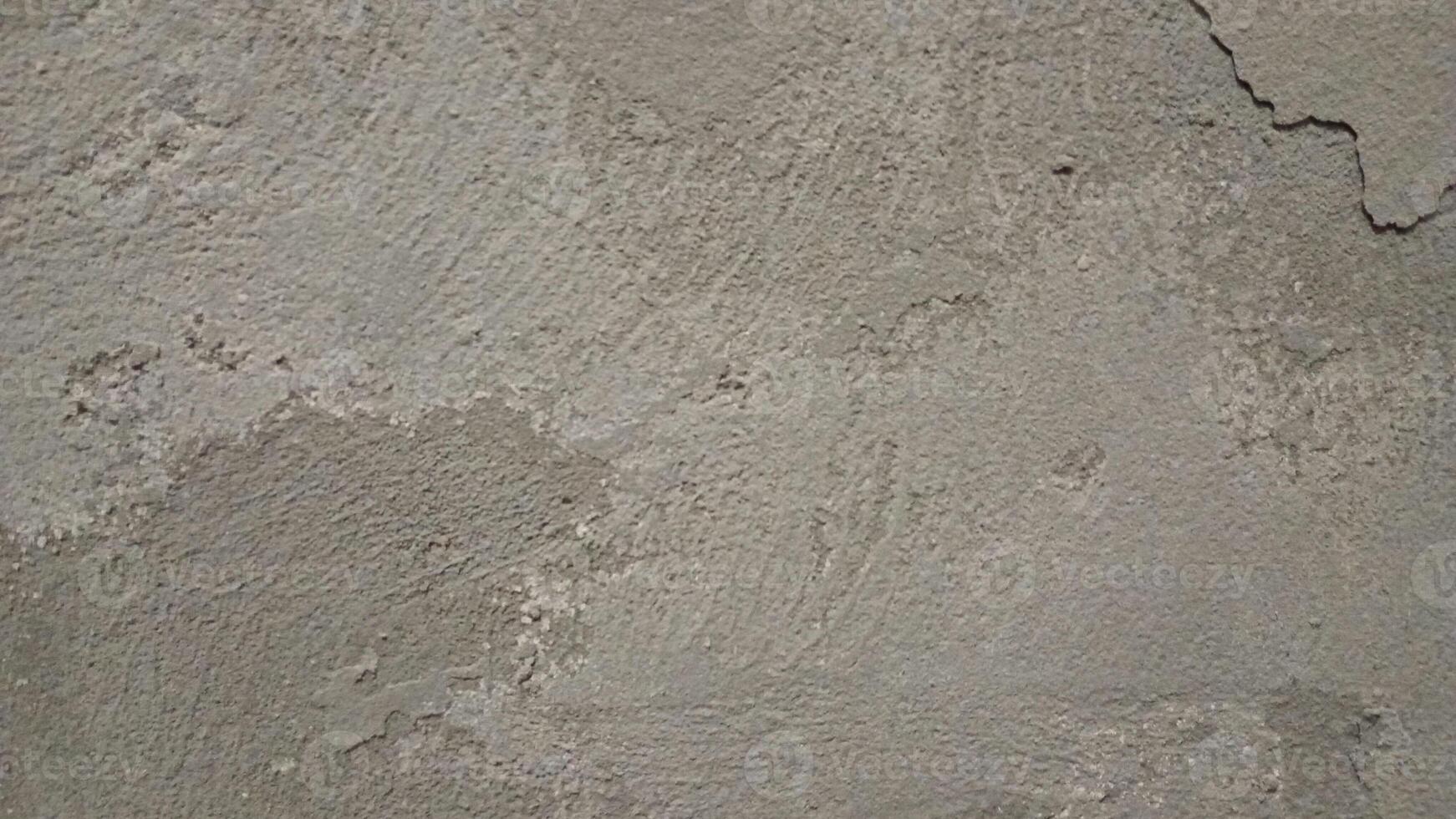 A close-up of a rough, worn-out concrete wall with a gritty texture, featuring cracks and crevices. The color is a dull, gray with subtle variations in shade, showing off the uneven surface. photo