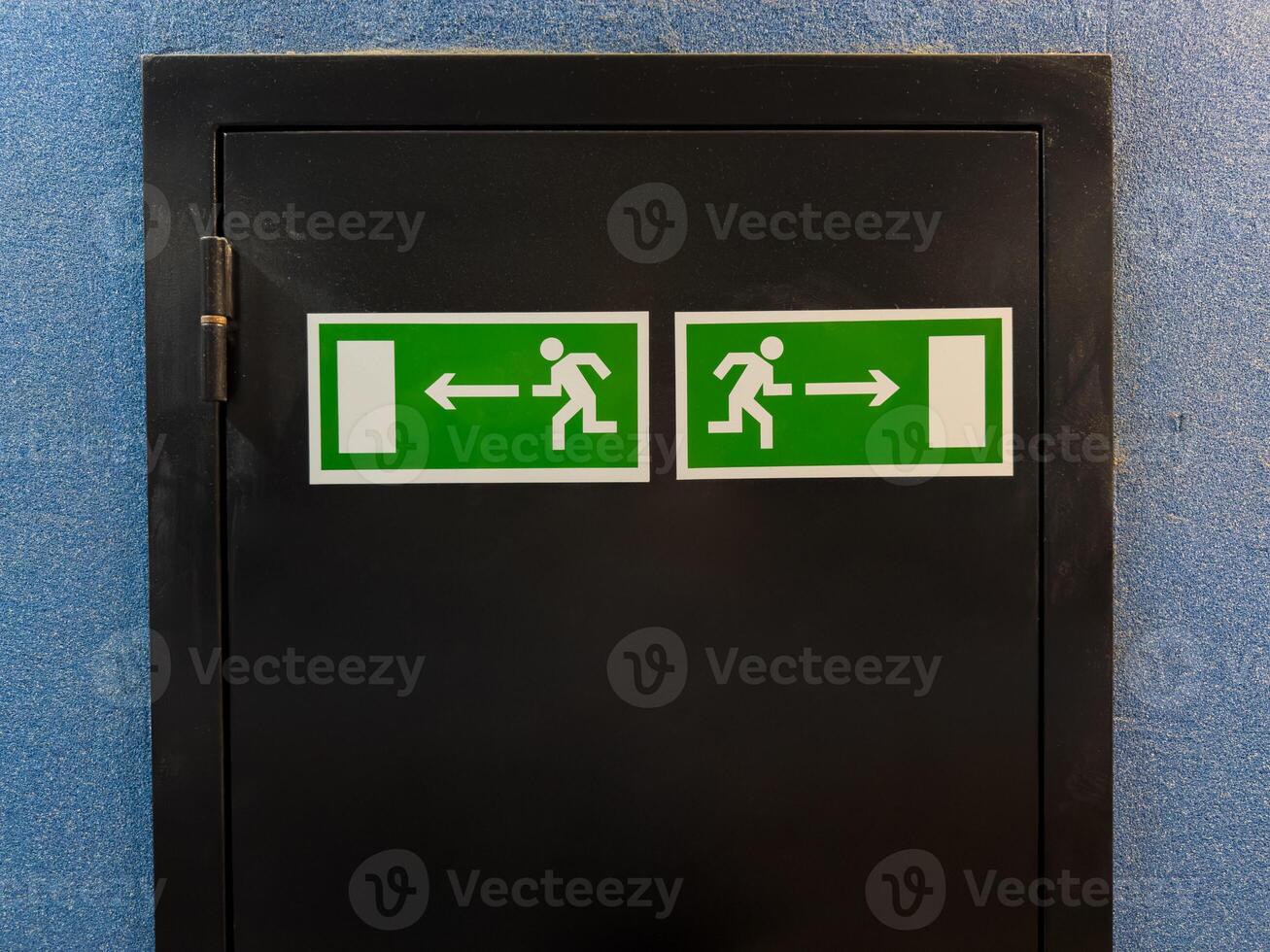 Two emergency exit signs with running figure and arrows pointing left and right placed on black door panel against blue wall, providing clear direction for safe evacuation in case of emergency photo