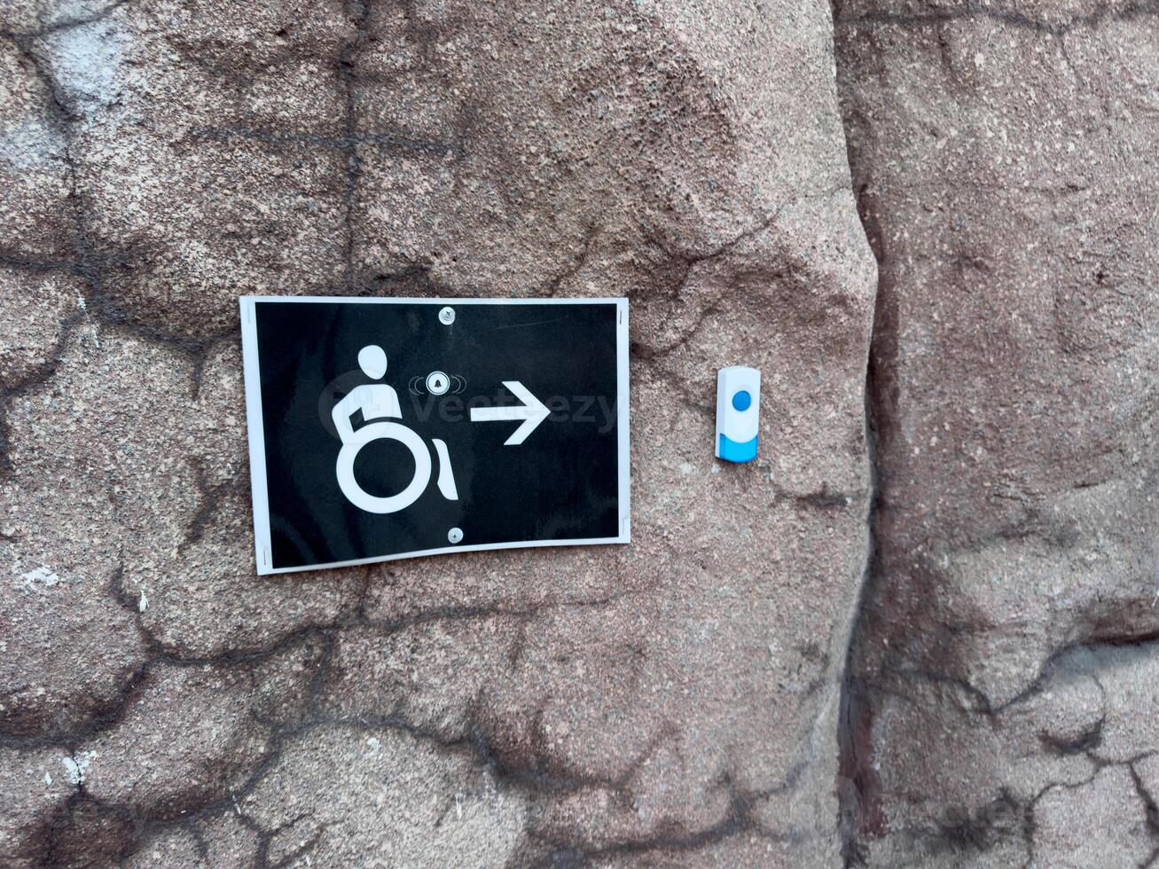 Accessibility sign with wheelchair icon and arrow mounted on textured stone wall next to service button, indicating direction and call support for people with limited mobility in public environment photo