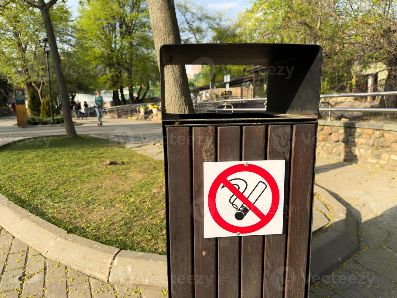 Close up of wooden outdoor trash bin with bold no smoking sign attached, placed in clean public park with trees on bright sunny spring day, promoting cleanliness photo