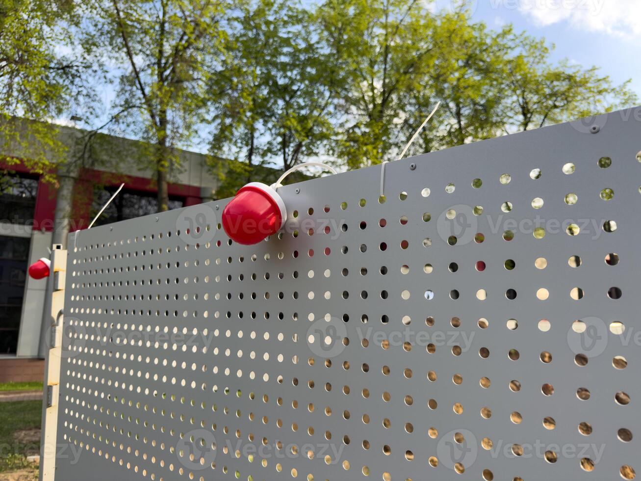 Modern perforated metal fence with red dome lights forms a sleek urban barrier near a contemporary public building. Functional design meets visual simplicity in this outdoor city infrastructure photo