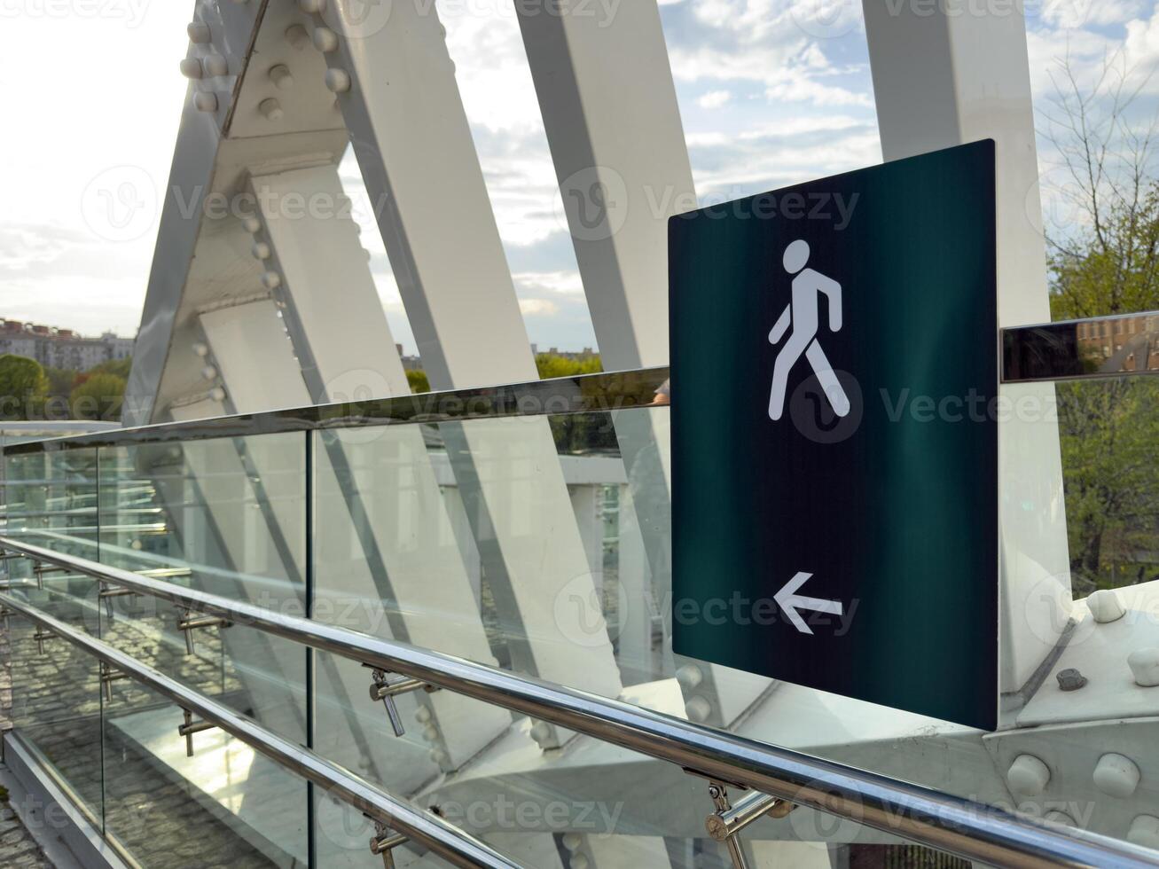 Close up of pedestrian walkway sign with walking figure and arrow on modern glass and steel bridge, with urban buildings and green trees visible in the background photo