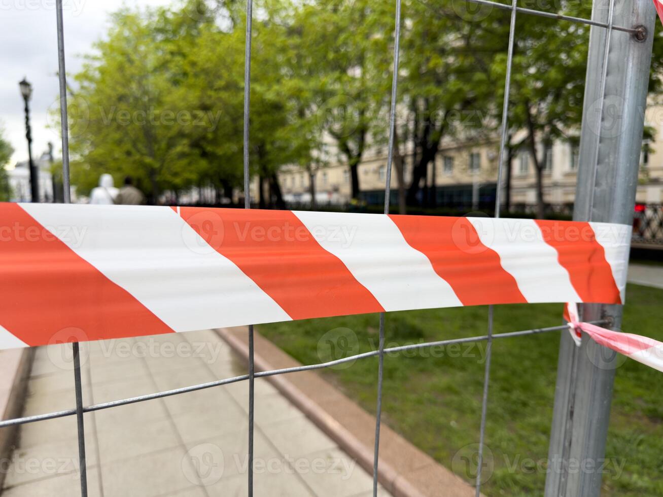 Red and white warning tape stretched across metal fence indicates restricted access in a public park area. Safety measure marks the boundary of an ongoing maintenance zone photo