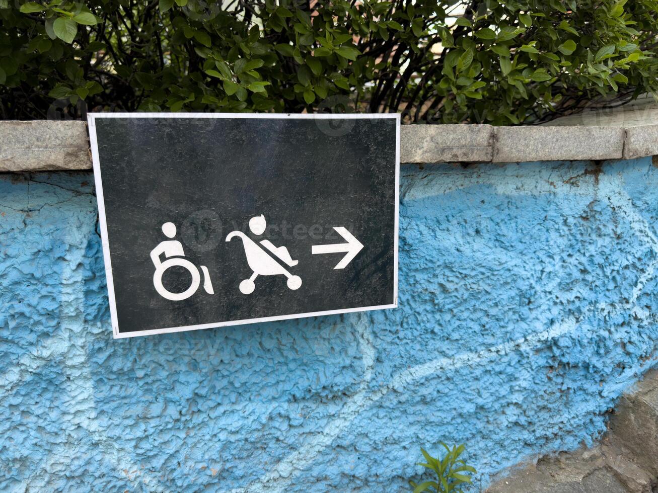 Closeup of black accessibility sign with white icons of wheelchair and baby stroller on textured blue wall, serving as clear directional symbol for inclusive access in public space or facility photo