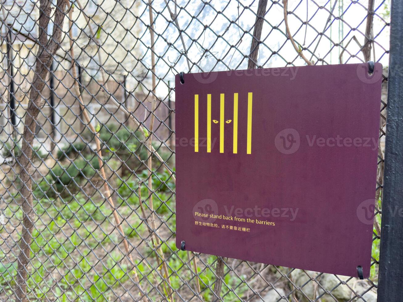 Zoo warning sign with yellow bars and eyes, suggesting dangerous animal behind the fence. Text advises visitors to stand back from the barriers for their safety and protection photo