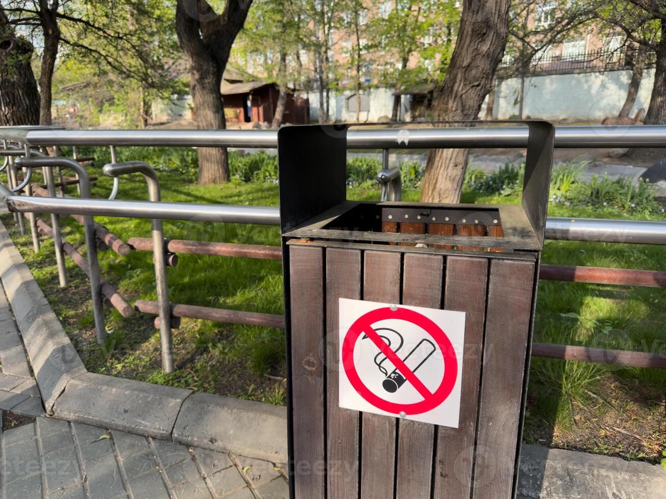 Outdoor wooden trash bin with no smoking sign, located in park area with metal railings and green grass on sunny day. Clean environment and public safety concept photo