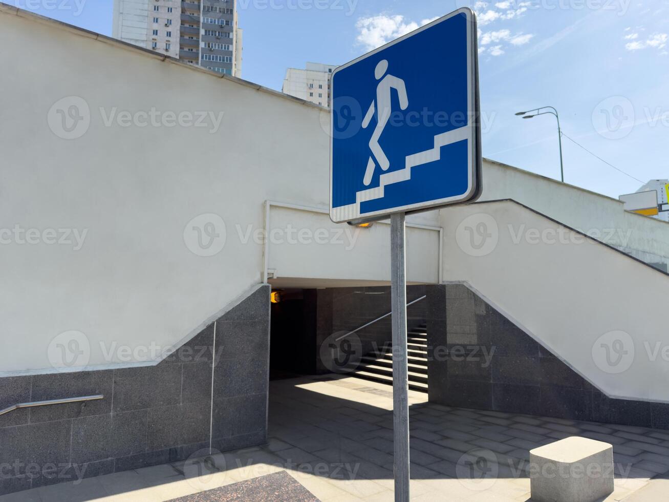Blue pedestrian sign showing figure descending stairs placed in front of underpass entrance with white wall and tile base, representing urban navigation and public infrastructure in city environment photo