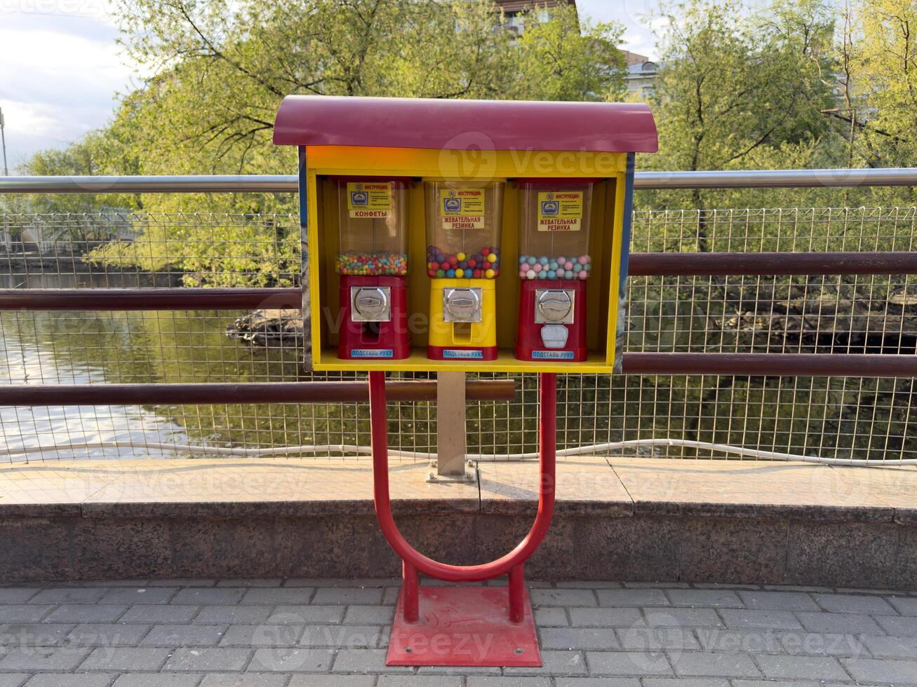 Colorful candy and gum vending machine stands on paved riverside path, inviting passersby to enjoy sweet treat. Surrounded by greenery, adds playful touch to the urban scene photo