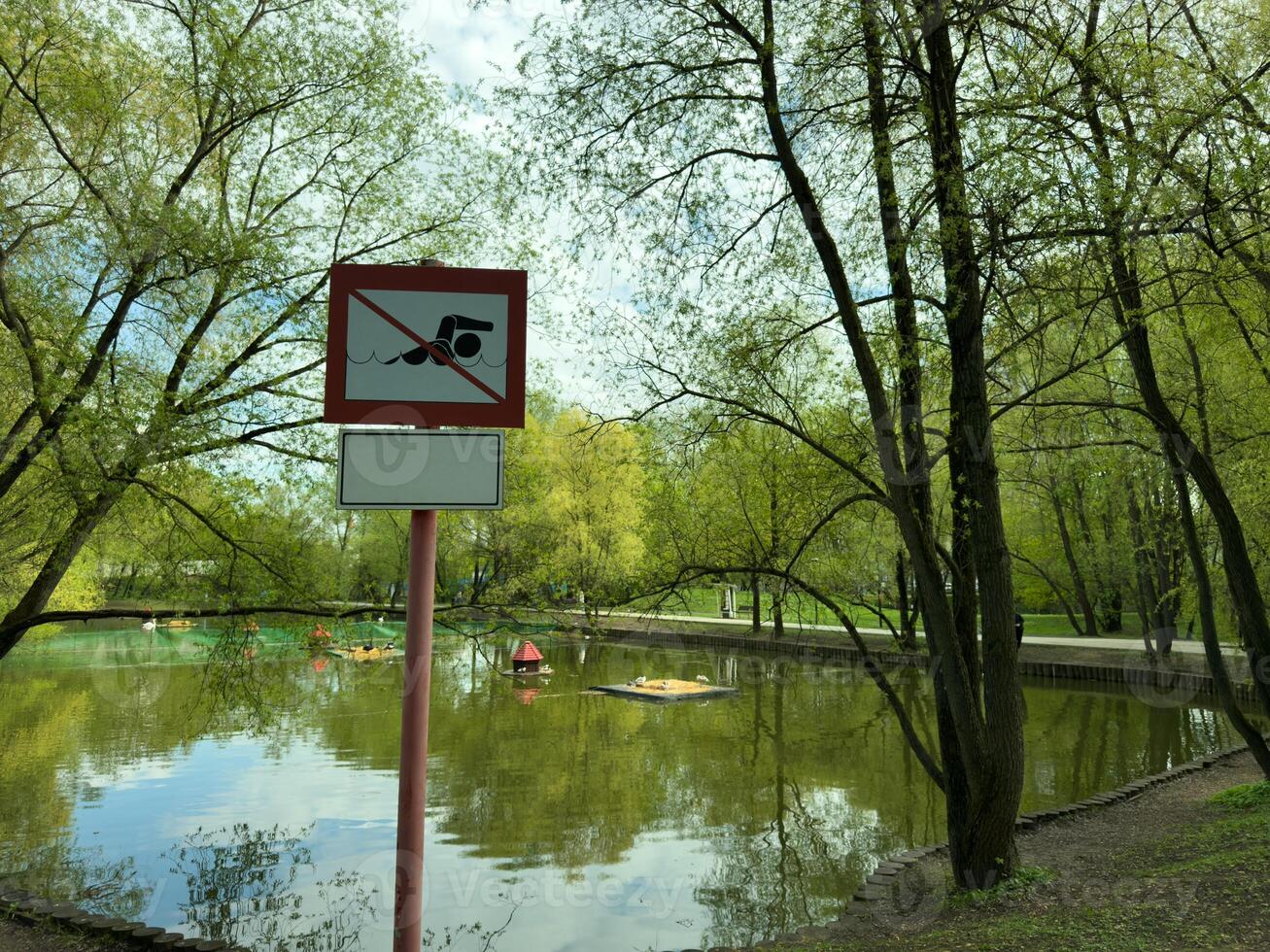 Warning sign by scenic pond in public park prohibiting swimming. The calm water reflects the surrounding trees, and area features pedal boats and small island for ducks photo