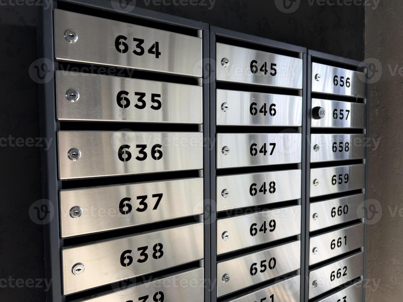 Row of modern stainless steel mailboxes with bold black numbers and built in locks mounted on dark wall in apartment building lobby, illustrating secure personal storage in urban residential space photo