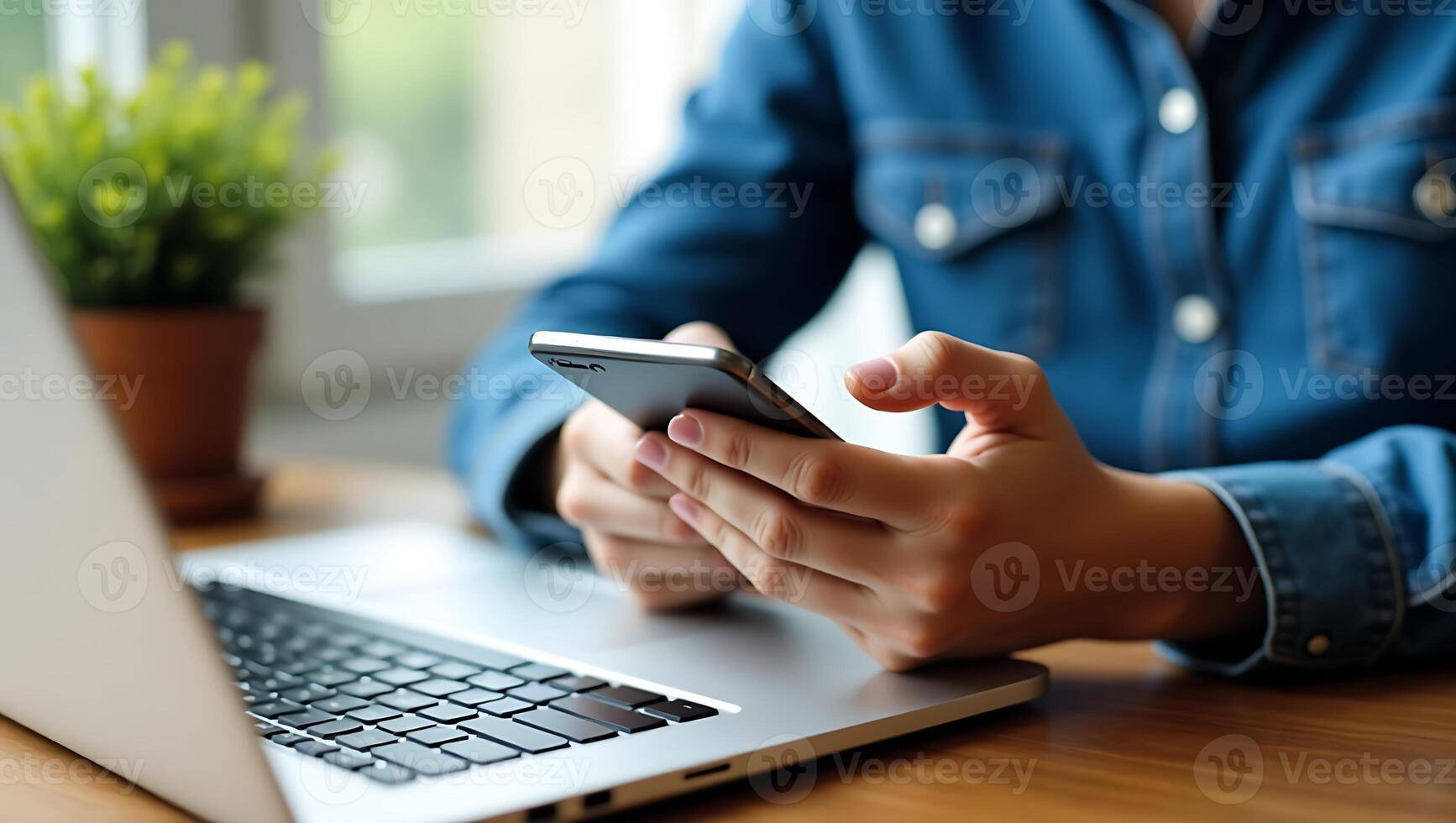 Woman using a smartphone near a laptop computer at a wooden table with a plant. photo