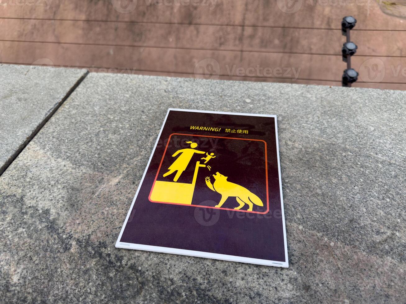 Caution sign with bright yellow pictogram warning about danger of lifting children near wild animals, placed on stone surface in zoo area, serving as visual safety reminder for visitors and families photo