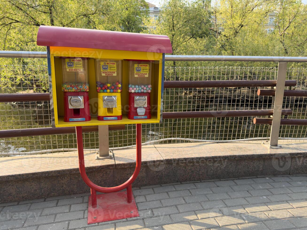 Bright vending machines with candy and chewing gum stand on city promenade by river. Cheerful colors contrast with greenery and calm water in background, evoking childhood memories photo
