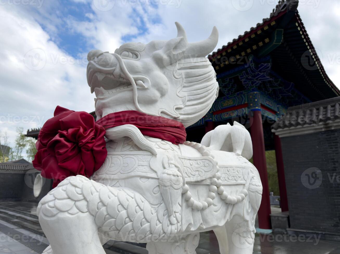 White stone dragon sculpture with decorative details and red ribbon in front of traditional Chinese architecture under cloudy sky, representing strength and cultural symbolism photo