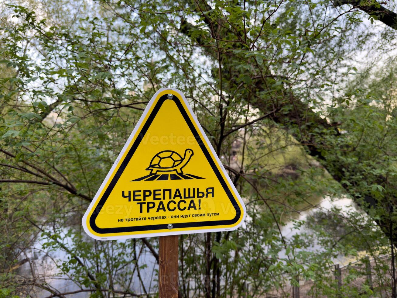 Yellow triangular turtle crossing sign in forest area near water with Russian text warning visitors not to disturb turtles, blending humorous tone with environmental awareness and wildlife protection photo