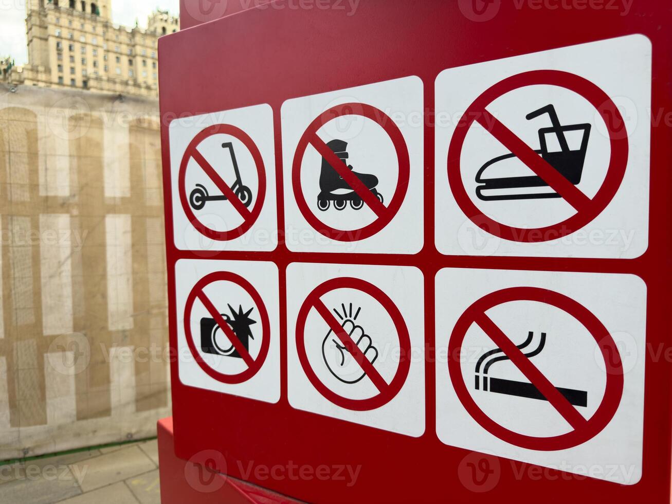 Set of bold red prohibition symbols on urban signboard illustrating restricted activities such as riding scooters, taking photos, smoking or making noise in public space near city building photo