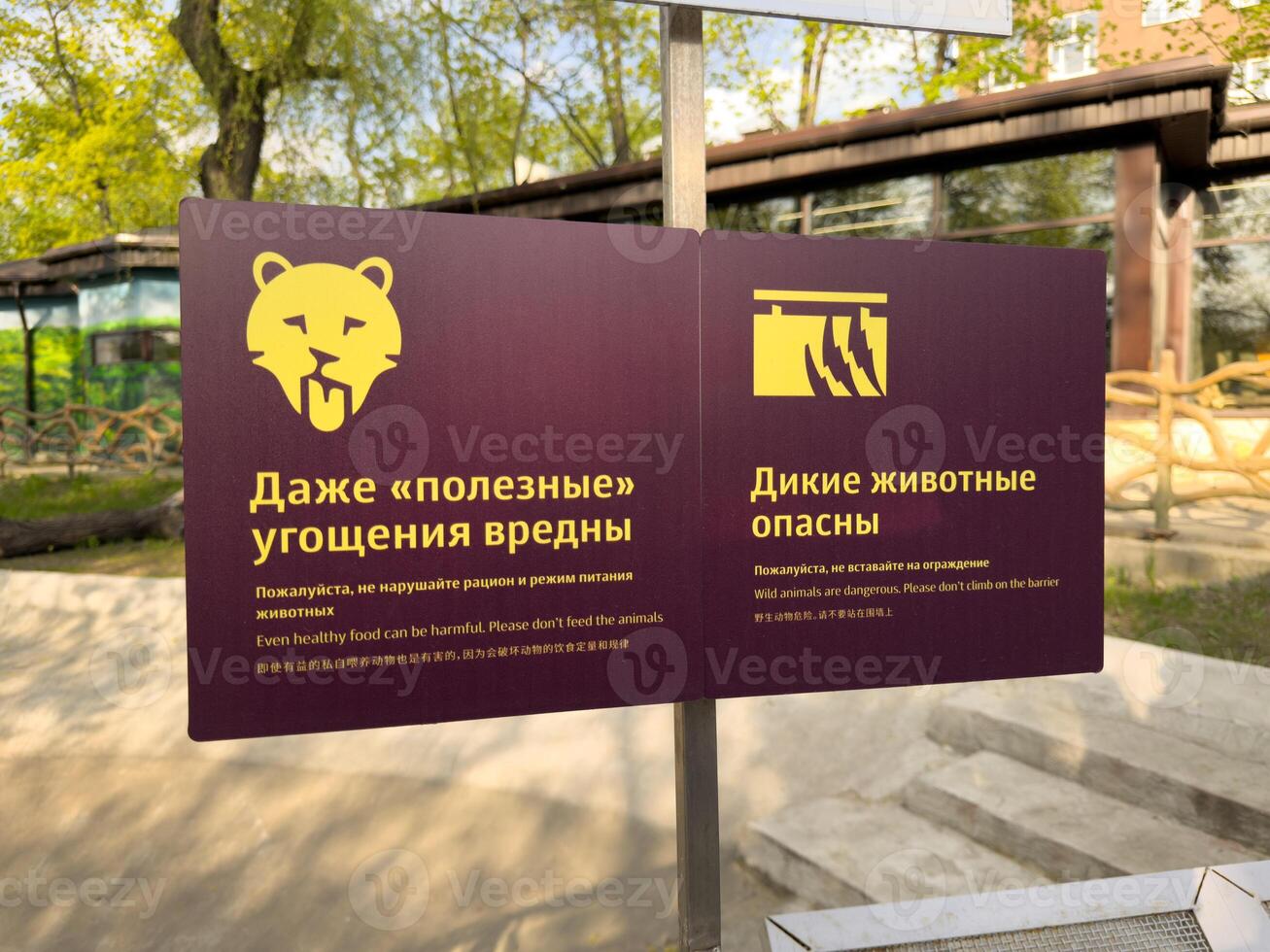 Warning signs in zoo with multilingual safety messages about feeding animals and climbing barriers, captured outdoors in natural daylight. Zoo sign in Russian with English and Chinese translations photo