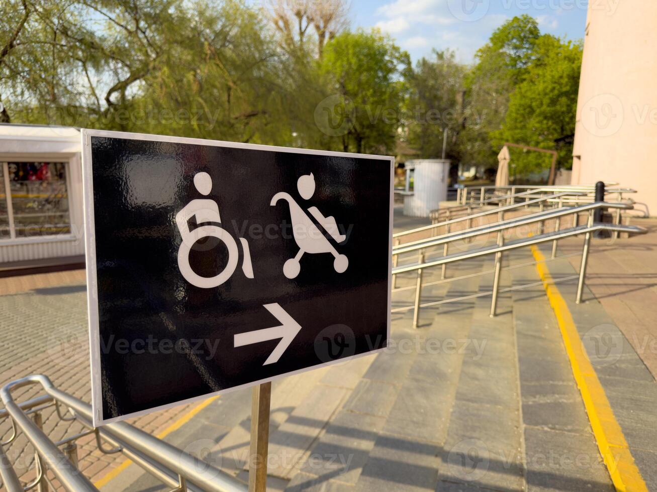 Accessibility sign showing direction to ramp for wheelchair users and strollers, located outside public building with handrails and trees in the background on sunny day photo