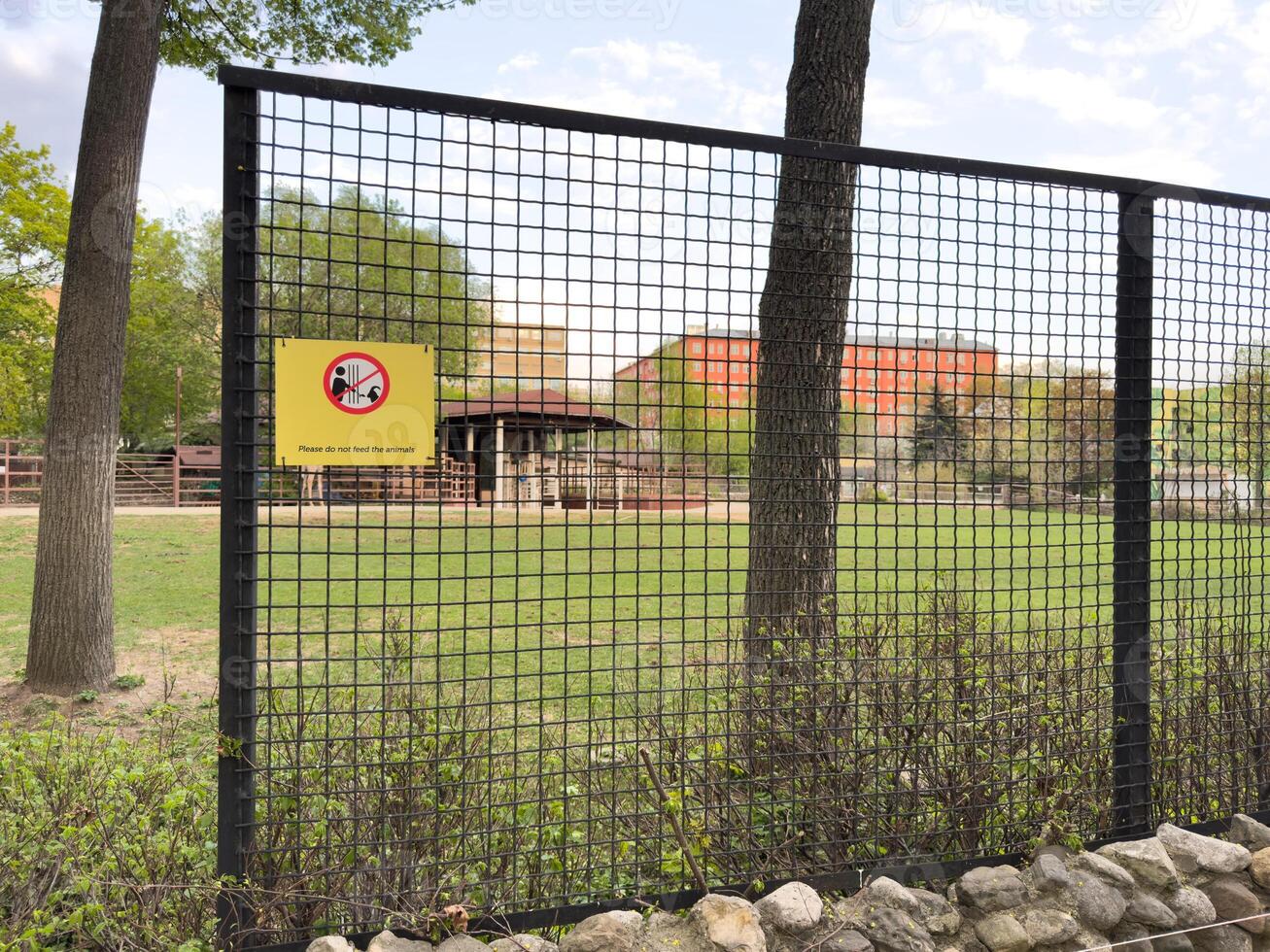 Wire fence with yellow warning sign in zoo area instructing visitors not to feed animals, surrounded by green grass, trees and enclosures, serving as protective barrier and rule enforcement element photo