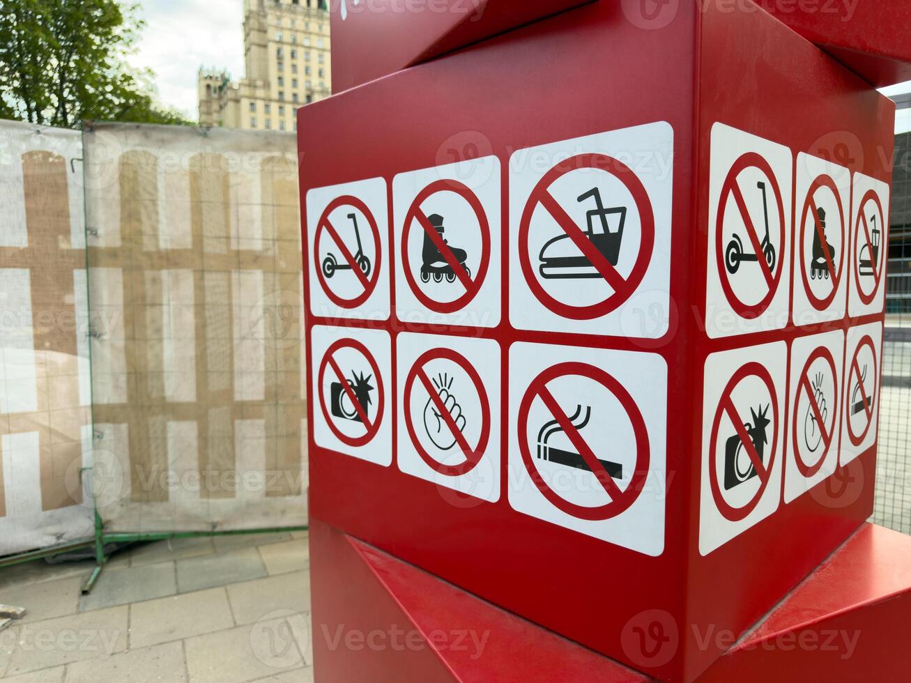 Red informational sign with six prohibition symbols against scooters, skating, bumper cars, photography, clapping, and smoking placed outdoors with blurred city background photo