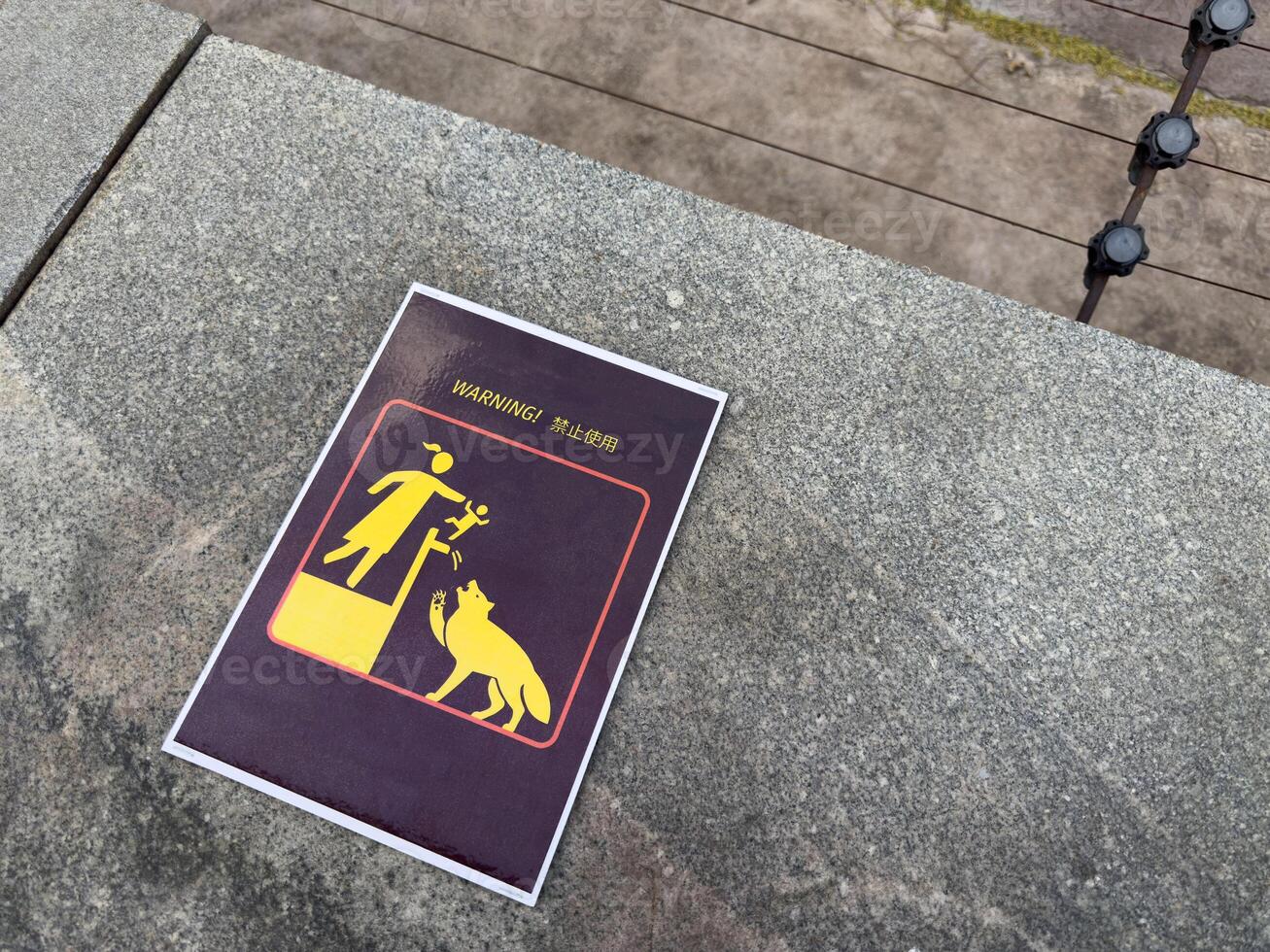 Warning sign with stylized graphic showing adult lifting child toward dangerous animal, placed on stone surface near electric fence in zoo, includes text in English and Chinese for visitors photo