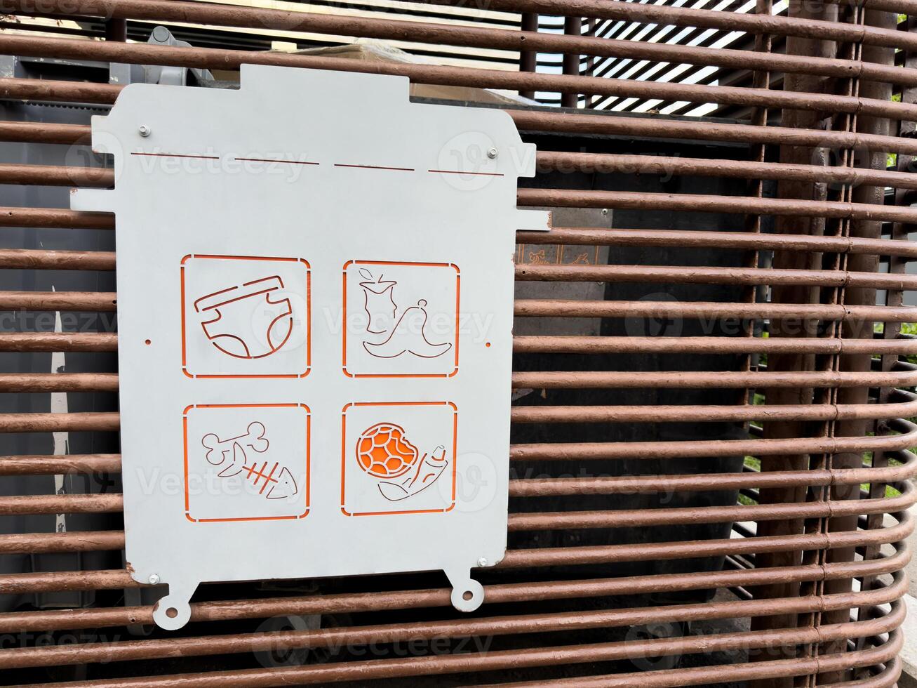Stylized waste bin sign with minimalist icons illustrating prohibited trash types. Modern metal plate design with orange line art symbols mounted on a metal slat surface in an urban setting photo