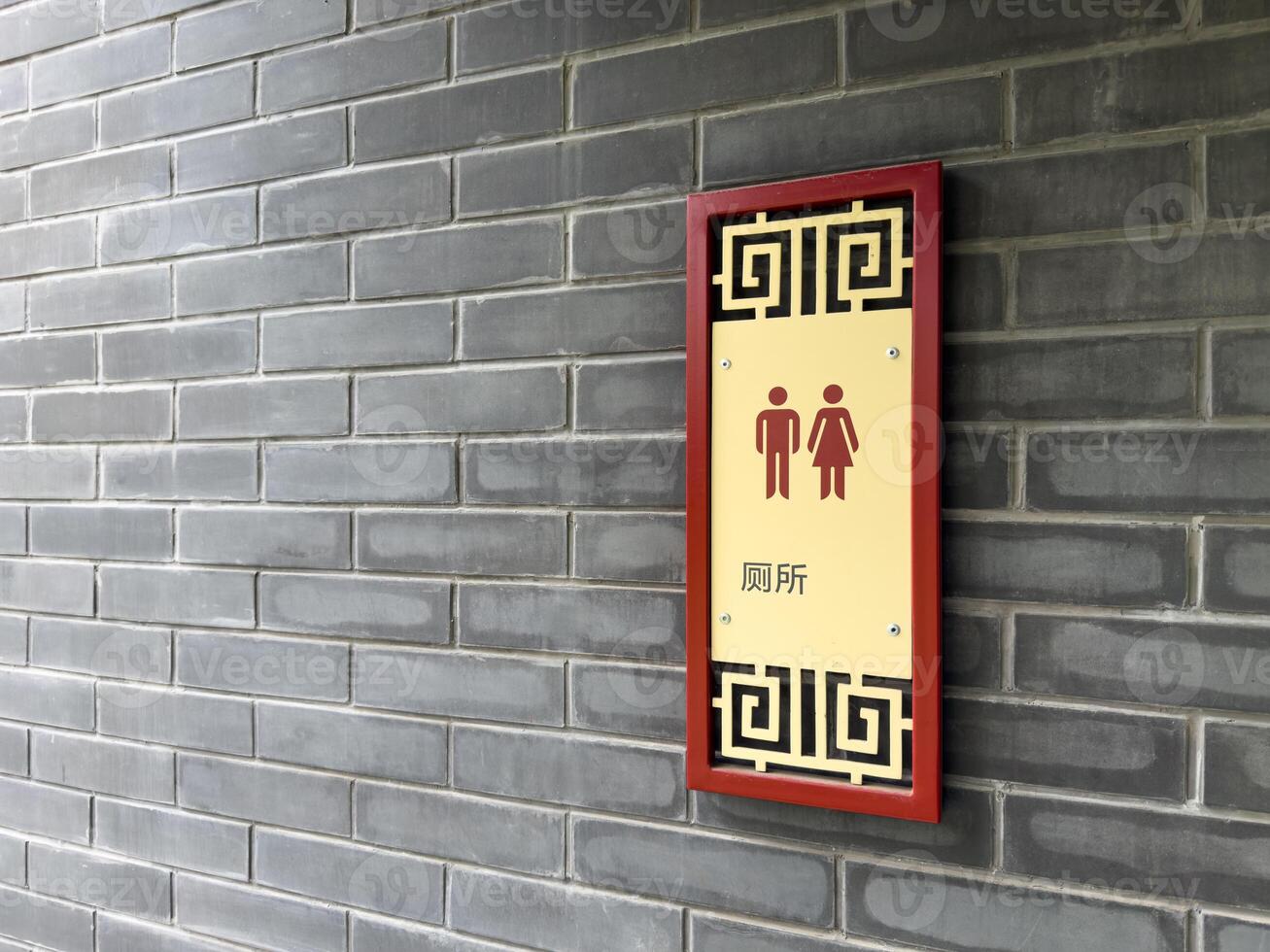 Wall mounted restroom sign with male and female pictograms and Chinese characters, framed with traditional Chinese ornament, attached to gray brick wall in an Asian inspired architectural setting photo