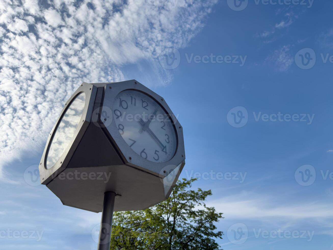 Modern outdoor clock mounted on tall metal pole with geometric casing and transparent panels, set against bright blue sky with scattered clouds in calm urban or park environment photo
