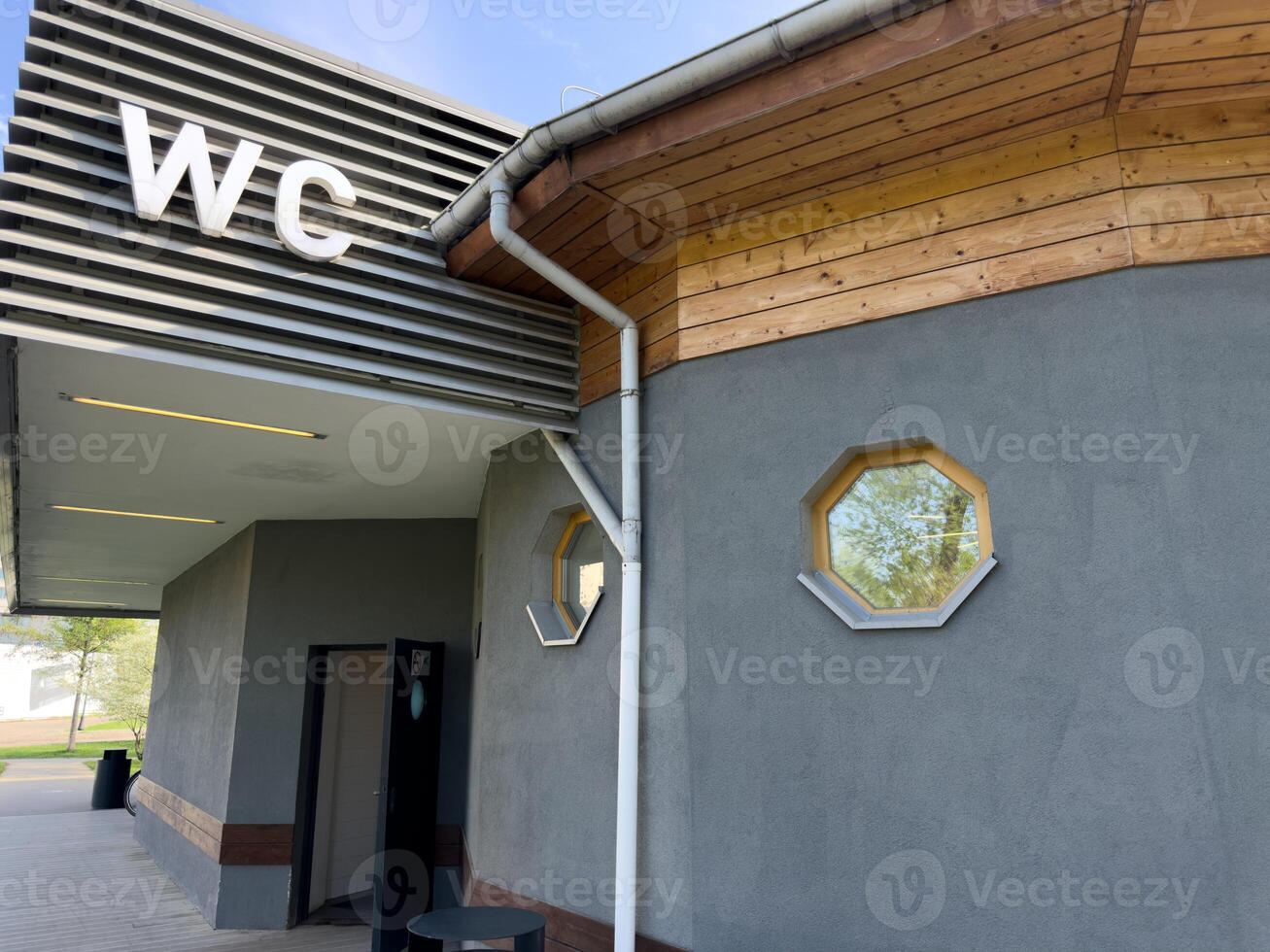 Contemporary public restroom with large WC sign, open door entrance, wooden roof detail and octagonal windows, combining practical accessibility with clean architectural elements in park environment photo