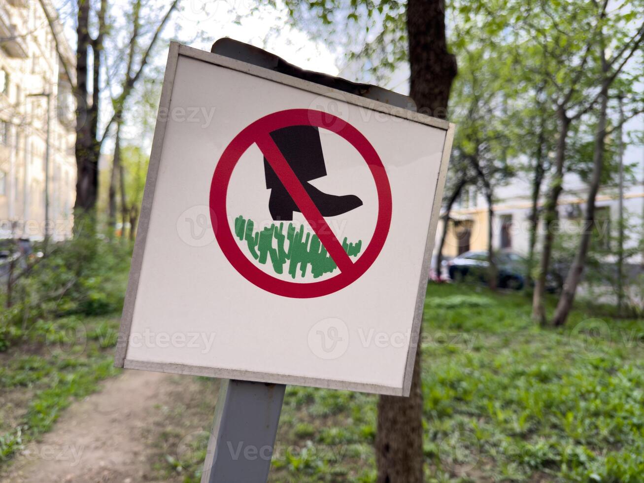 Warning sign in urban park prohibiting walking on the grass. Signpost with red circle and black boot pictogram placed in a green public area surrounded by trees and buildings photo