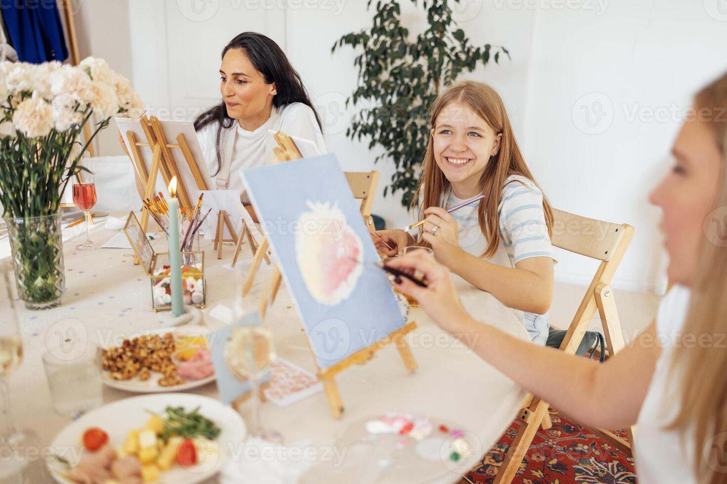 A young focused woman in a white T-shirt and apron paints a picture at a table and an easel. photo