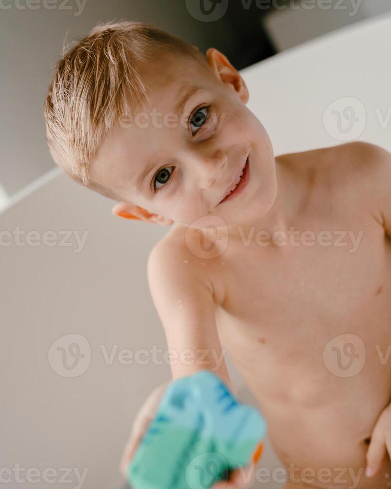 A playful 4-year-old boy sits in a bright white bathtub, his hands busy scooping and pouring water with toy cups, while sunlight streams through the windows, filling the room with warmth photo