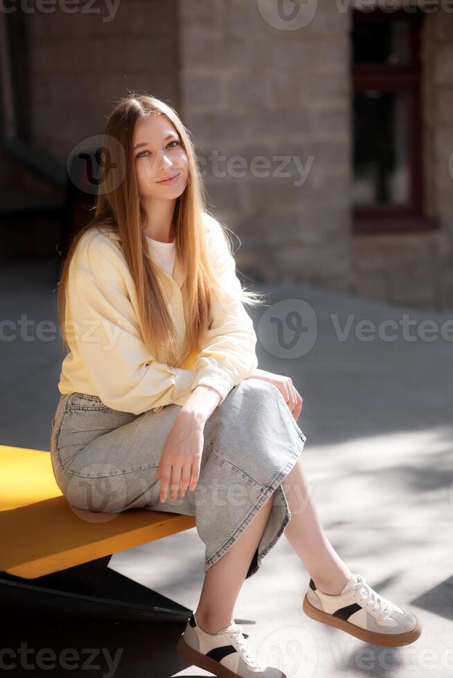 Young woman sitting on a yellow bench in sunlight wearing casual attire with a relaxed expression in an urban setting photo