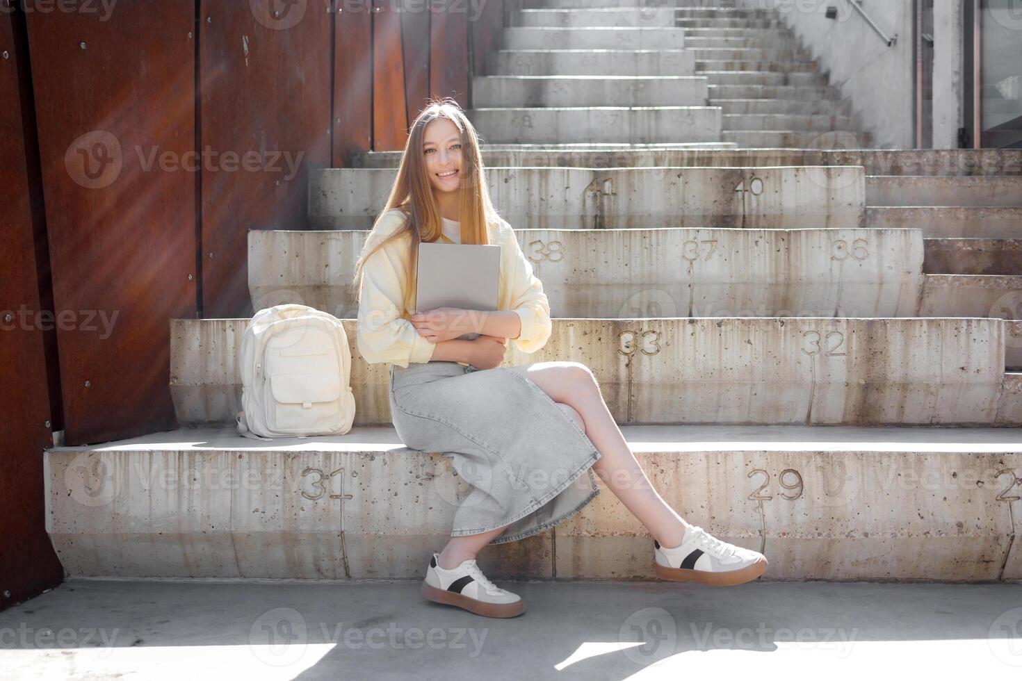 Young woman sitting on concrete stairs in a modern urban setting, preparing for studies with a laptop and backpack photo
