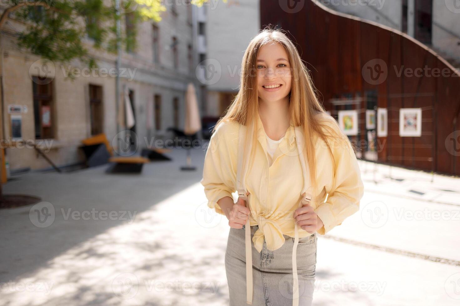 Young woman smiling in sunlit courtyard while wearing casual outfit and posing for camera during afternoon photo