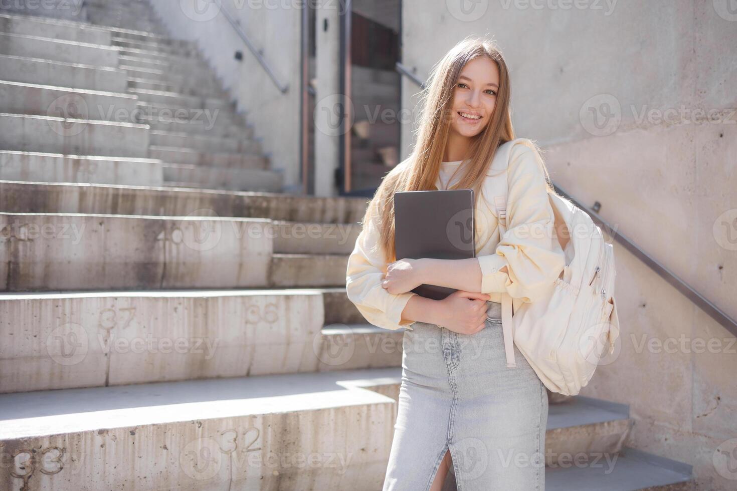 Student poses on outdoor stairs, smiling while holding laptop, showcasing a modern academic setting and casual style photo