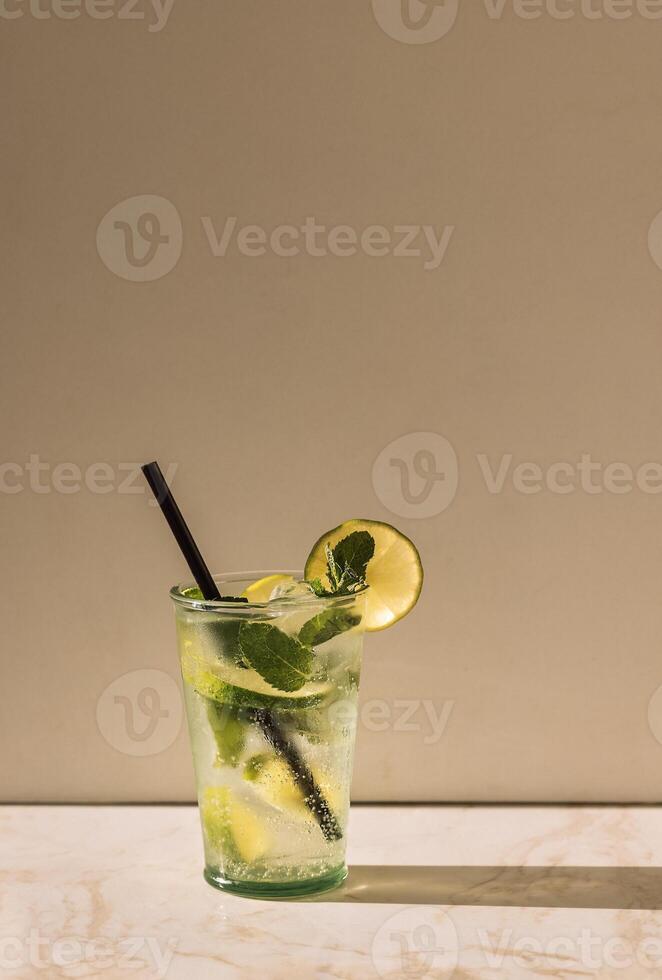 Close-up of a glass of delicious mojito with lime and fresh mint. Tasty lemonade with ice cubes on a marble table. A cup of cool citrus cocktail and a straw. photo