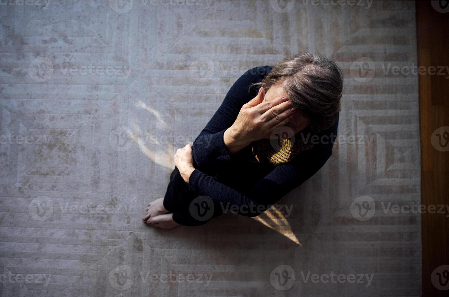 An elderly woman suffers from a headache at home. A mature lady is sitting on the floor and holding her forehead with her hand. View from above. photo