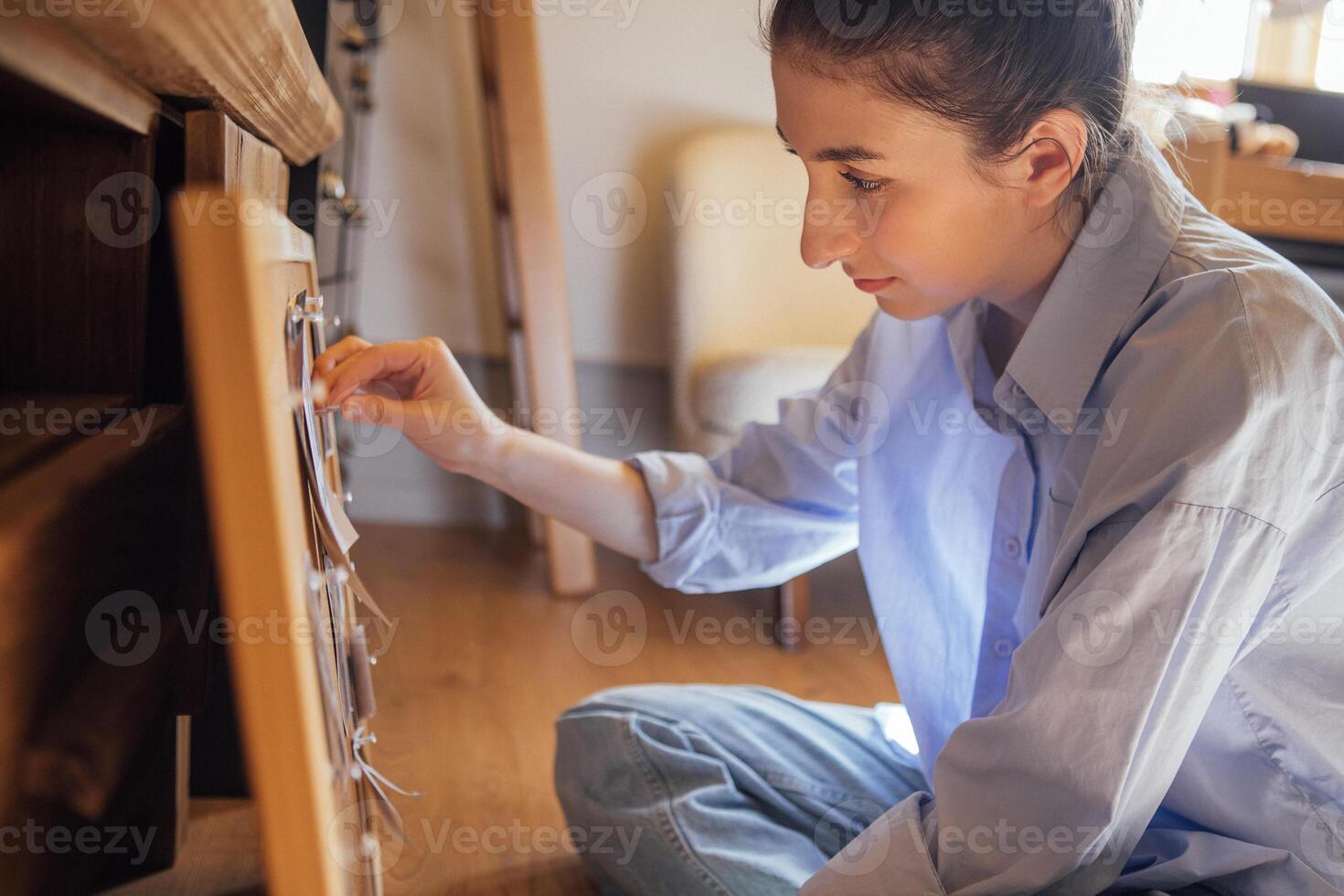 Close-up of a young woman in casual clothes sitting on the floor in front of a cork board. The girl pins the photos to the board.