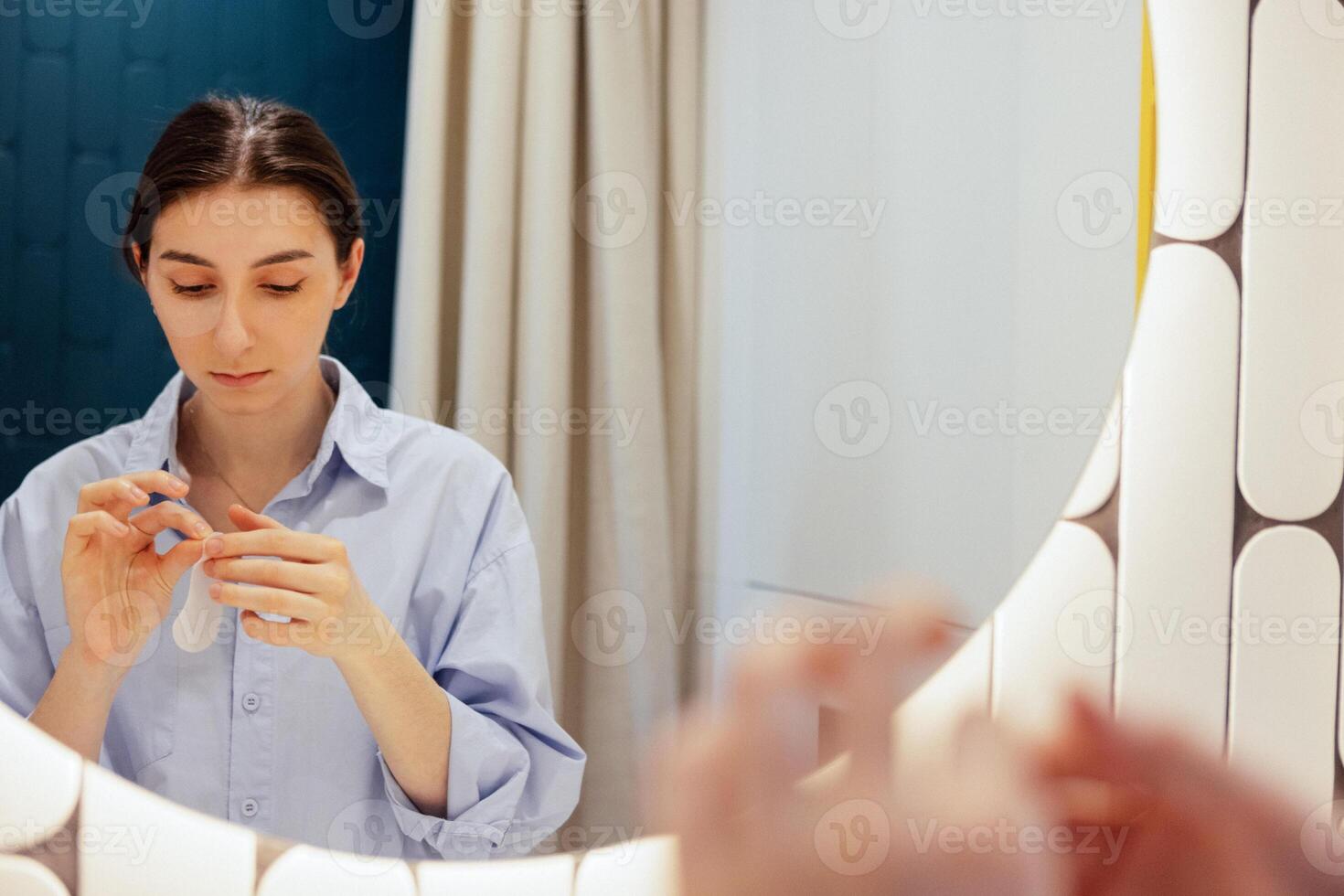 A young woman is applying moisturizing patches under her eyes in the bathroom. The reflection of a cute girl holding patches in her hands in a mirror. photo
