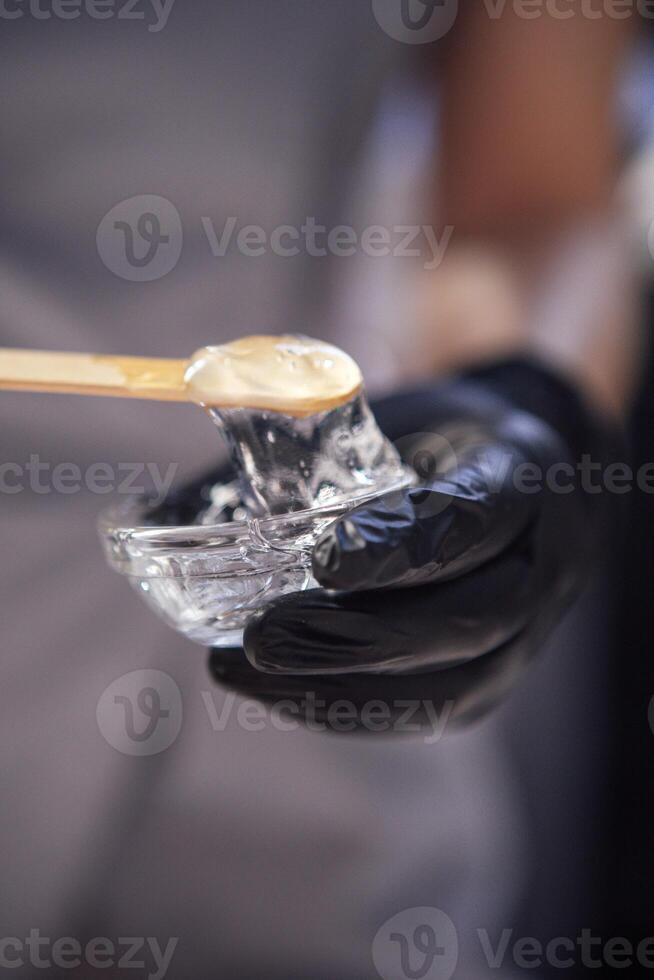 Close-up of a cosmetologist hands with a glass bowl and moisturizing gel. The dermatologist hands in black gloves hold a jar of nourishing serum and a wooden spatula. photo