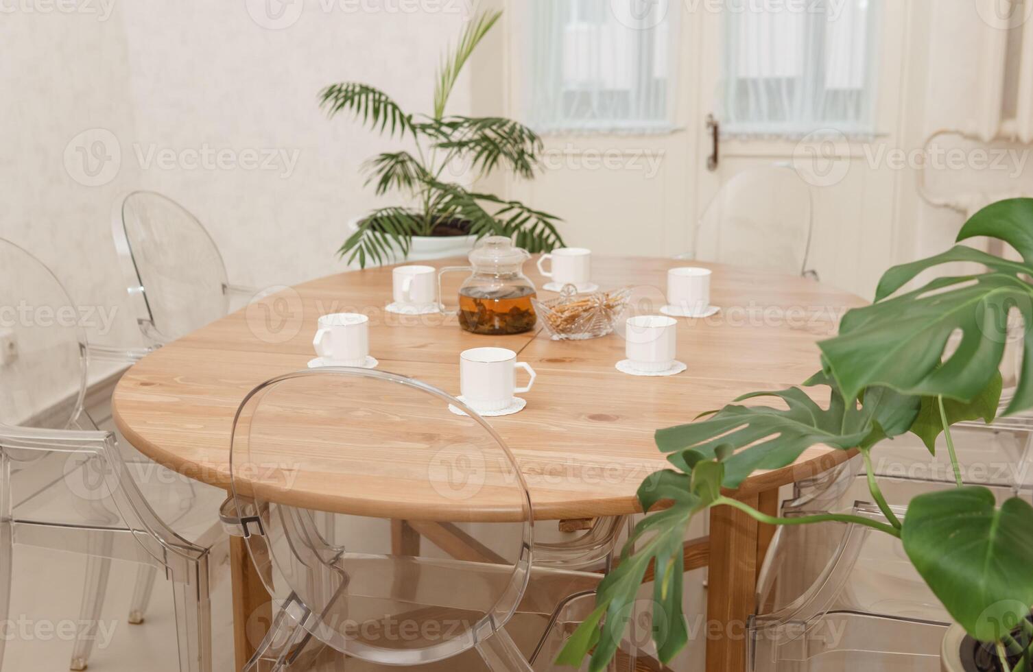 Cozy dining setting featuring round wooden table surrounded by transparent chairs, complemented by lush green potted plants, creating a warm and inviting interior atmosphere photo