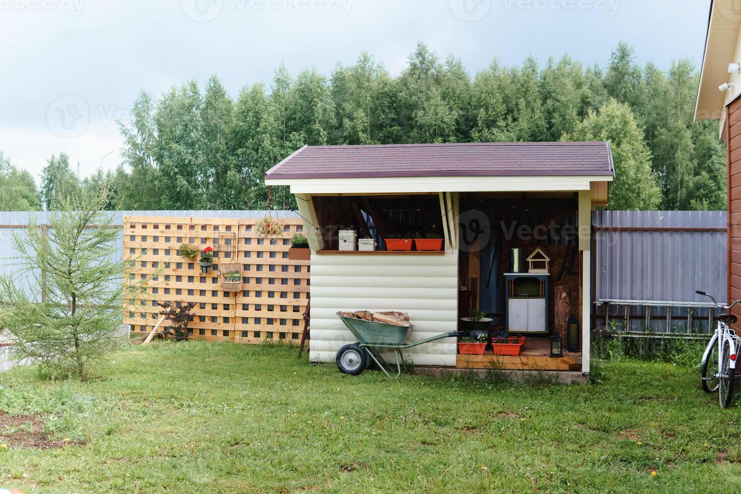 A rustic storage shed stands within a backyard adorned with a wheelbarrow overflowing with wood, a decorative wooden lattice, and potted plants, ideal for illustrating rural life. photo