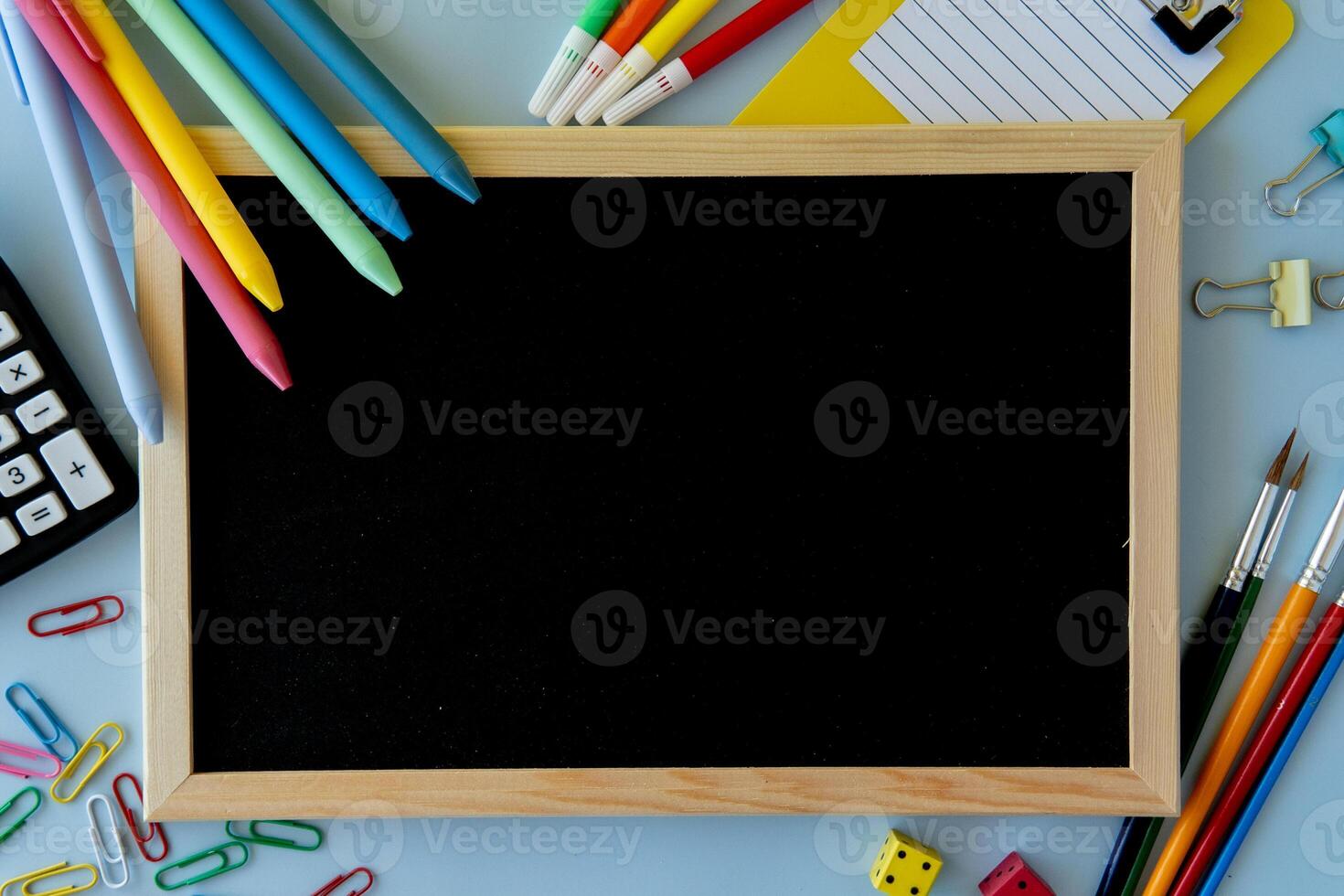 Top view of school desk with blackboard vibrant pens paper clips binder clips calculator paintbrushes colorful dice and clipboard on blue surface concept for education creativity and student planning photo