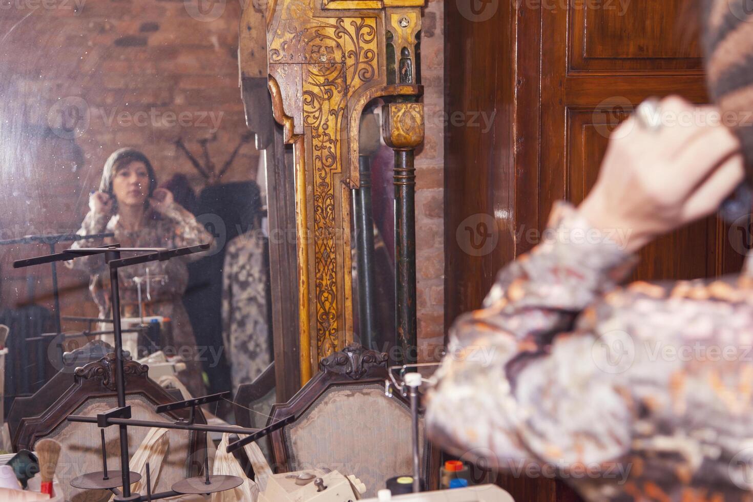 a young model tries on a handmade hat while looking at herself in a large mirror in a tailor's shop photo