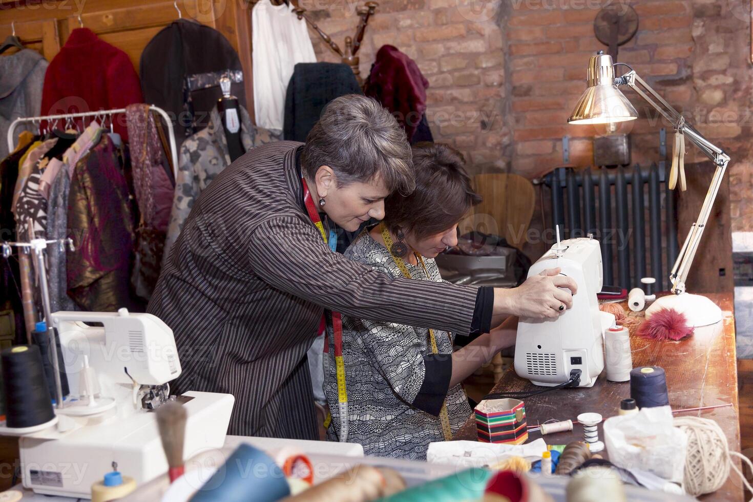 mature seamstress teaching sewing machine work to an apprentice in a fashion atelier photo